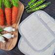 Two natural color hemp potholders partially stacked on top of one another. Ribbing/edging on pot holders is a grey color.  Top potholder you can see the loop for hanging.  Also pictured with a wooden cutting board, fake green onions, carrots and snap peas.