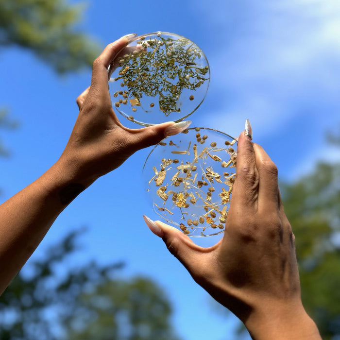 Two round resin coasters held to the sky by mark skin hands with long fingernails.  Top coaster is clear resin filled with hemp flower.  Bottom coaster is clear resin coaster filled with hemp baste fiber.