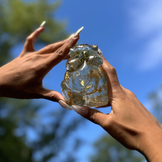 Two hands with long nails holding a hand poured resin figurine of a full bodied woman, with hemp buds at heart-center and gold flake throughout.  She is holding the figurine in the air with blue sky in the background. 