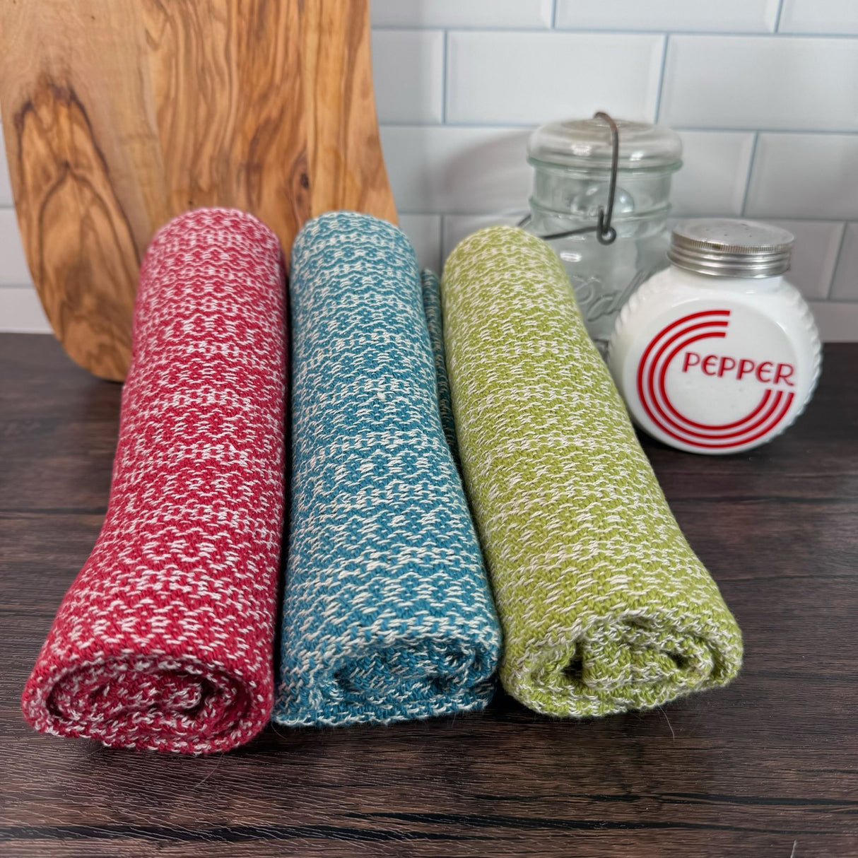 Three hemp blend dish towels (red/white, teal/white, lime green / white) rolled up individually on a counter.  In the background there is a cutting board, pepper shaker and mason jar.