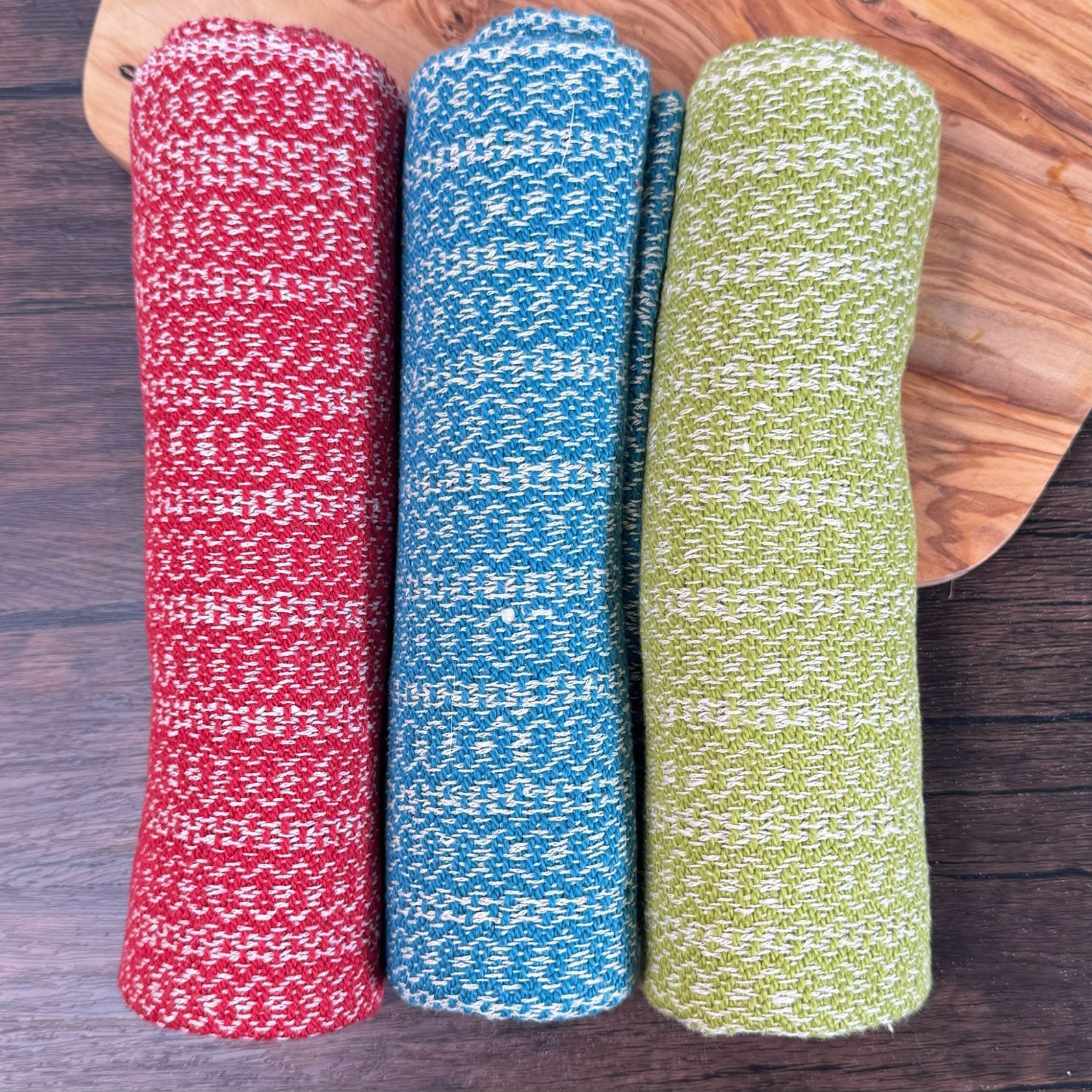 Three hemp blend dish towels (red/white, teal/white, lime green / white) rolled up individually on a counter.  In the background there is a wooden cutting board.  Picture is a top down view of the rolled towels.