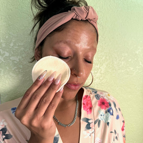 Closeup face view of a model wearing a floral lightweight bathrobe is using a hemp facial round to clean her face.  Model is also wearing large hoop earrings and a pink headband.