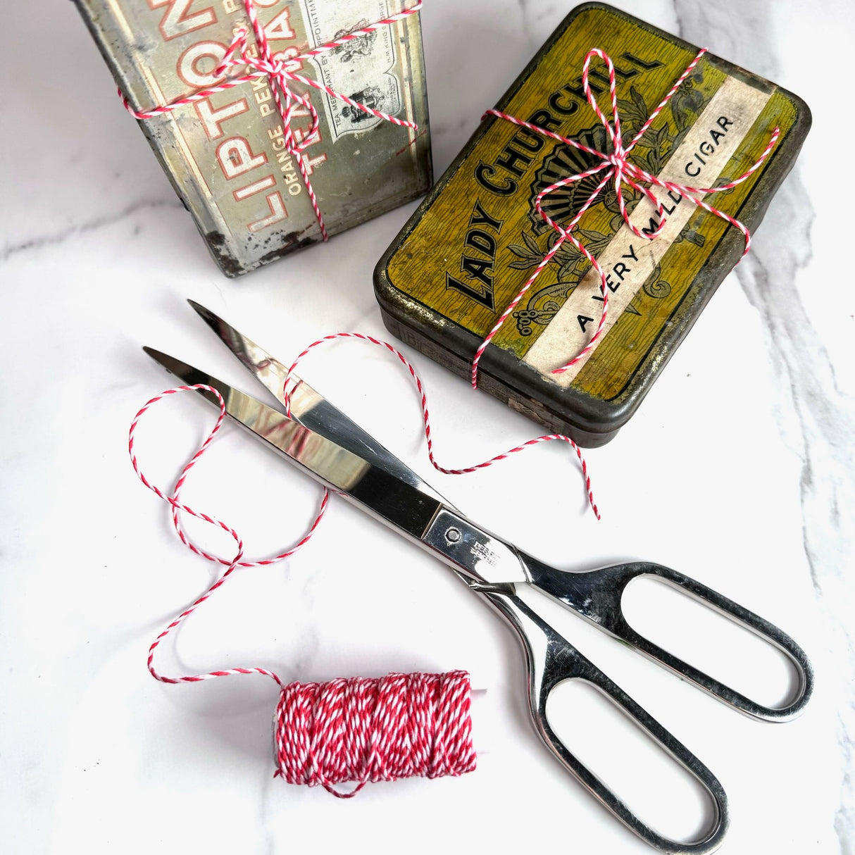 Red and white spiral hemp cord partially unwound on a white marble table.  Pictured with classic style silver scissors and two antique tin boxes with the hemp cord tied around them as if it's ribbon on a package. 