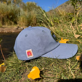 Side view of a Blue hemp vote baseball hat with a pencil or joint holder on the side that says "root for hemp."  Pictured outside on green grass with some yellow leaves and a river in the background. 