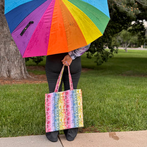 Person holding a rainbow umbrella and matching tote bag outdoors.
