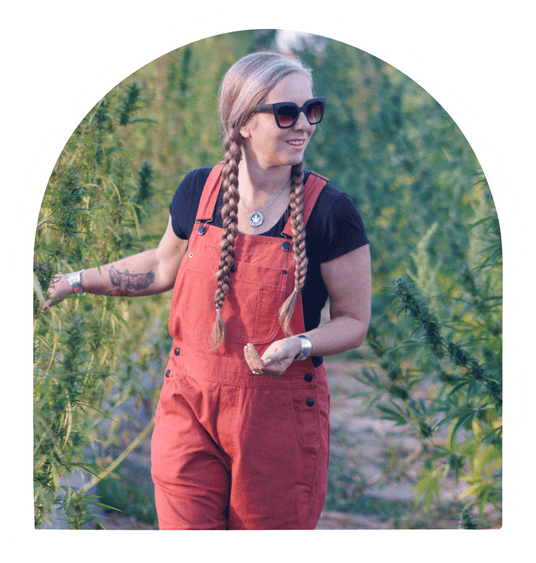 Picture of Lauren Gibbs on a hemp farm, wearing red hemp overalls and a black tee shirt and touching the hemp plants. 