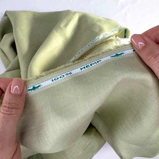 Green fabric with a visible '100% Hemp' label held by hands on a white background