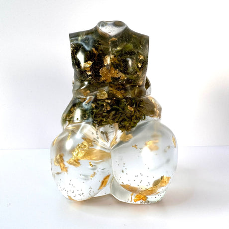 Closeup rear view of a hand poured resin figurine of a full bodied woman, with hemp buds at heart-center and gold flake throughout.  Figurine is sitting on a white surface with a white background.