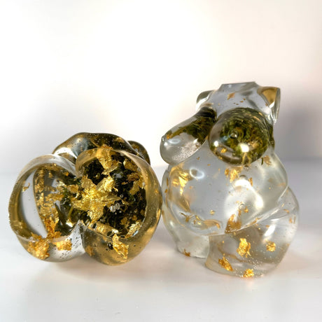 Two hand poured resin figurine of a full bodied woman - one is the front view and another is the bottom view. 
 Both are with hemp buds at heart-center and gold flake throughout to show the variation in the art. Scene is on a white surface with a white background.