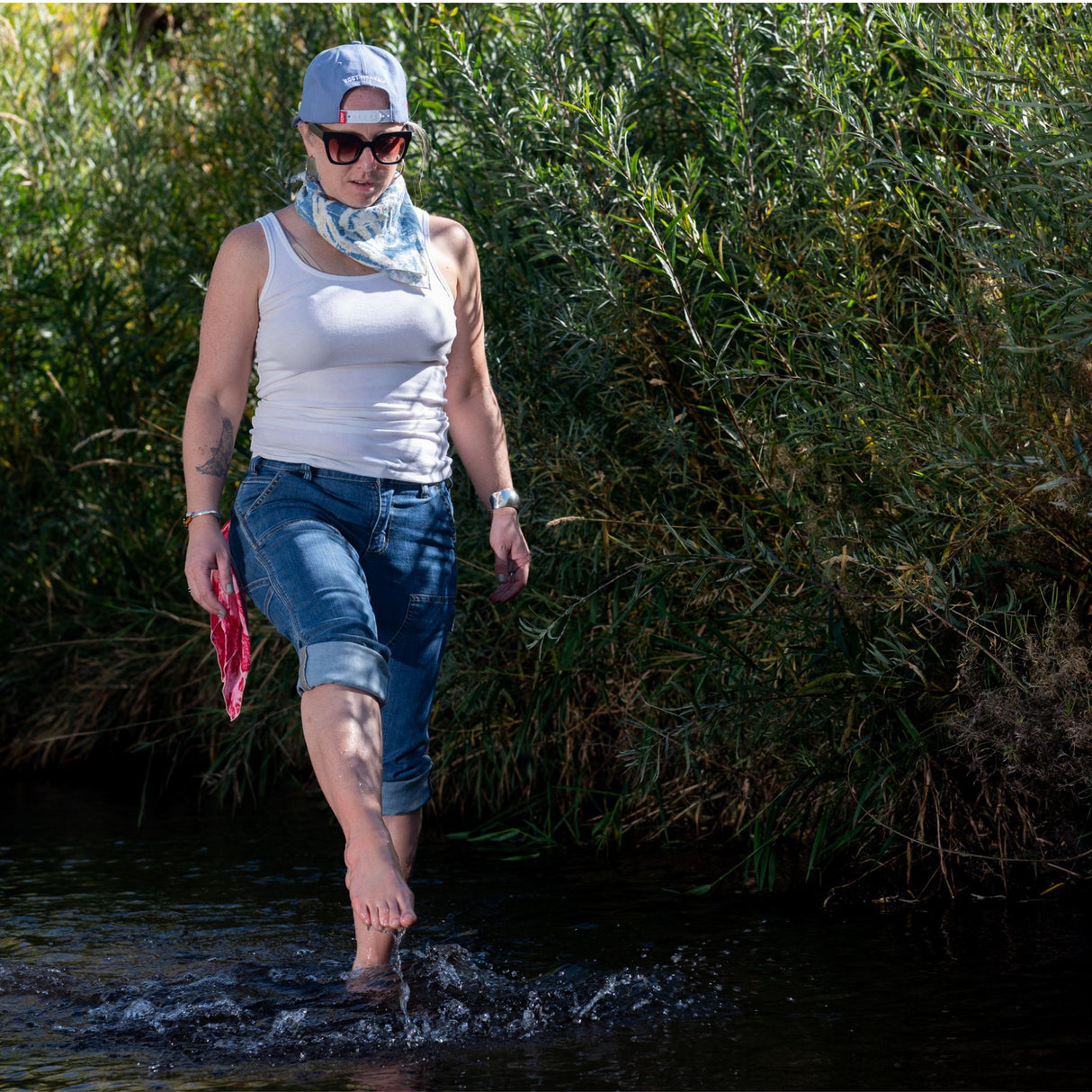 Woman walking through a creek in bare feet - she is wearing rolled up blue jeans and a white tank top.  She also has a red hemp bandana hanging from her back pocket and a blue hemp bandana wrapped around her neck.  She is wearing black sunglasses and a backwards baseball cap. 