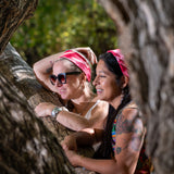 Two women leaning against a large tree branch, both wearing red hemp bandanas around their head.  Both girls are smiling and looking at something that is out of view of the scene. 