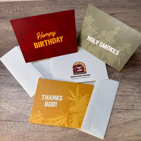 Folded standing red hemp paper greeting card with the words "hempy birthday." Folded standing green greeting card with the words "holy smokes." Folded laying down flat yellow greeting cards with the words "thanks bud." Also picture is the back of the greeting card with the root for hemp words and logo along with words that read "Printed on tree-free hemp"