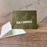 Green hemp paper greeting card with white words that read "holy smokes." Graphic on front of card includes hemp plant underlying the words and in light green.  Pictured with an unsealed hemp paper envelope.