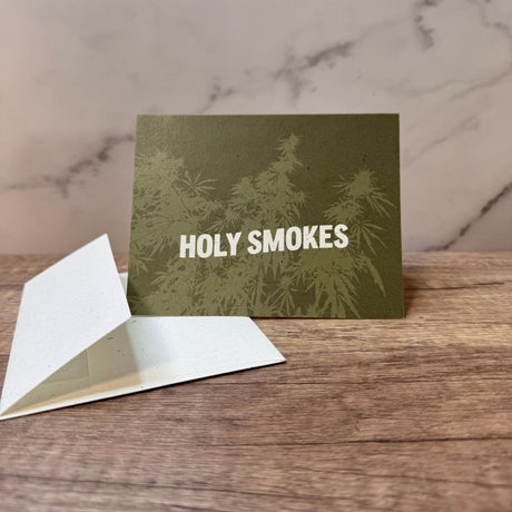 Green hemp paper greeting card with white words that read "holy smokes." Graphic on front of card includes hemp plant underlying the words and in light green.  Pictured with an unsealed hemp paper envelope.