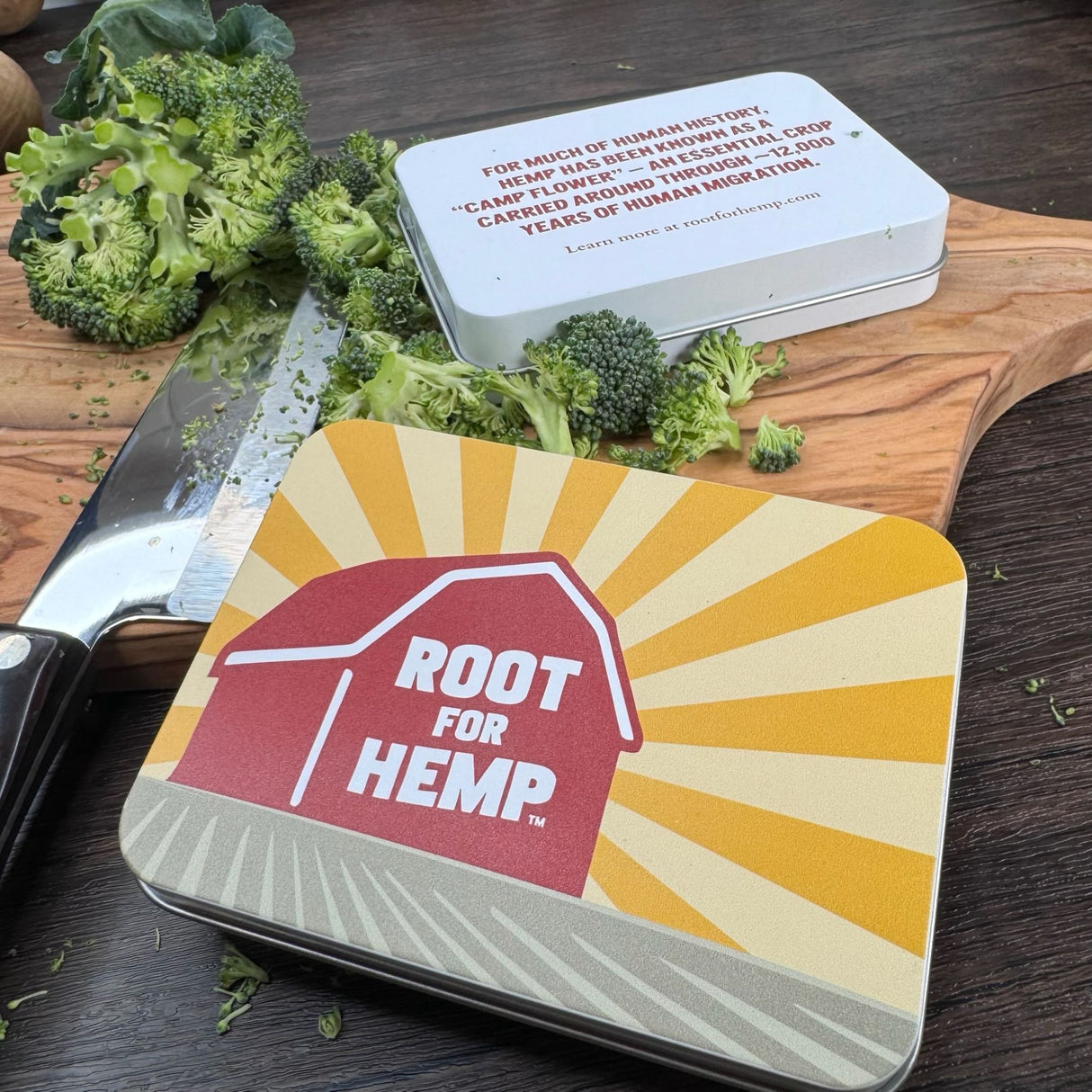 Front of pocket tin - red barn with words "root for hemp", yellow sun rays, and green field.  Back of tin with hemp educational message.  Also pictured with a wooden cutting board, knife, and chopped broccoli.