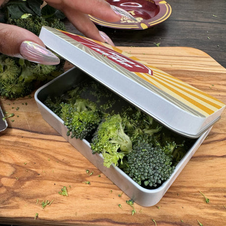 Partially open view of a pocket tin filled with broccoli.  Tin is white on the sides, silver on the inside.  Dark skin hand is holding the tin open.  Also pictured with a wooden cutting board and an ash tray.