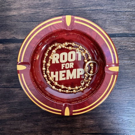 Round metal ash tray with red and gold with dark red leaf pattern, "Root for Hemp" on center of dish, show as being used as a jewelry tray - holding a ring and a necklace.