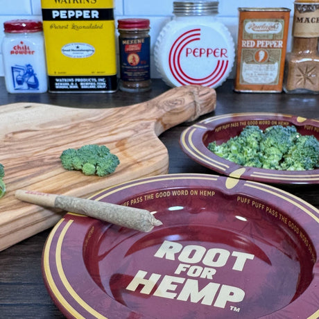 Closeup view of inside of the ash tray with the words "Root for Hemp" on center of dish, "puff puff pass the good word on Hemp" on the inside rim. Two ash trays pictured with a wooden cutting board, broccoli and a variety of vintage pepper jars.