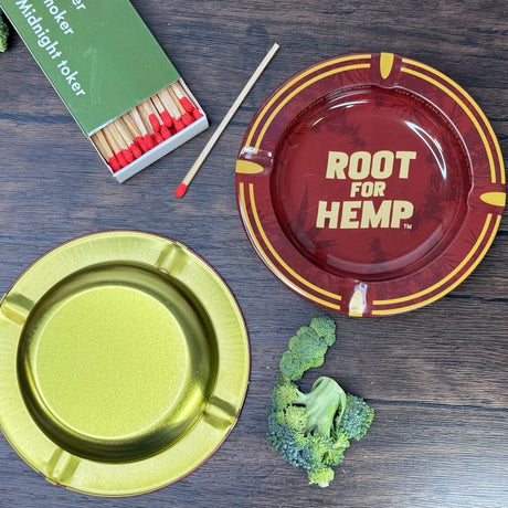 Bottom viewof the ash tray - bottom is gold.  Top view of the ash tray  which Red and gold with dark red leaf pattern, "Root for Hemp".  Also pictured with a book of matches and chopped broccoli.