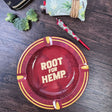 Round metal ash tray with red and gold with dark red leaf pattern, "Root for Hemp" on center of dish, "puff puff pass the good word on Hemp" on the inside rim. Background has a toker poker with hemp leaves, broccoli and a joint. 