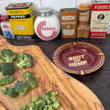 Round metal ashtray with 5.5 inch diameter pictured with vintage kitchen spice canisters and chopped broccoli on a cutting board. Red Ash Tray has 'Root for Hemp’ printed on base in gold font and 'puff puff pass the good word on hemp' imprinted on inside rim in gold font.