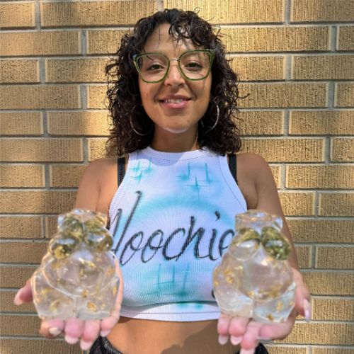 Profile picture of Stassi holding two pachamama resin products, wearing a white tank top with the words hoochie, green glasses, large hoop earings.