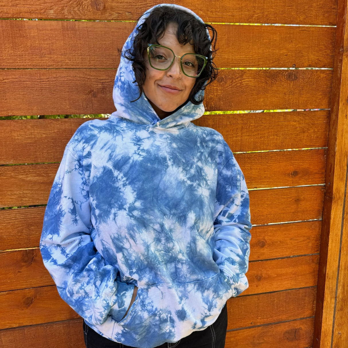 Front view of a model wearing an indigo tie dye hoodie with a the words.  She has the hood up over her head.  She is wearing green glasses, black pants, and has a wooden fence in the background. 