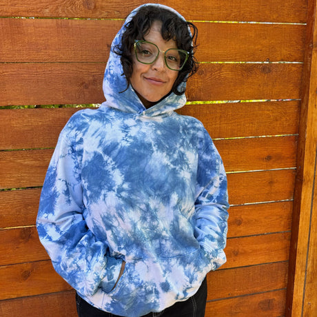 Front view of a model wearing an indigo tie dye hoodie with a the words.  She has the hood up over her head.  She is wearing green glasses, black pants, and has a wooden fence in the background. 