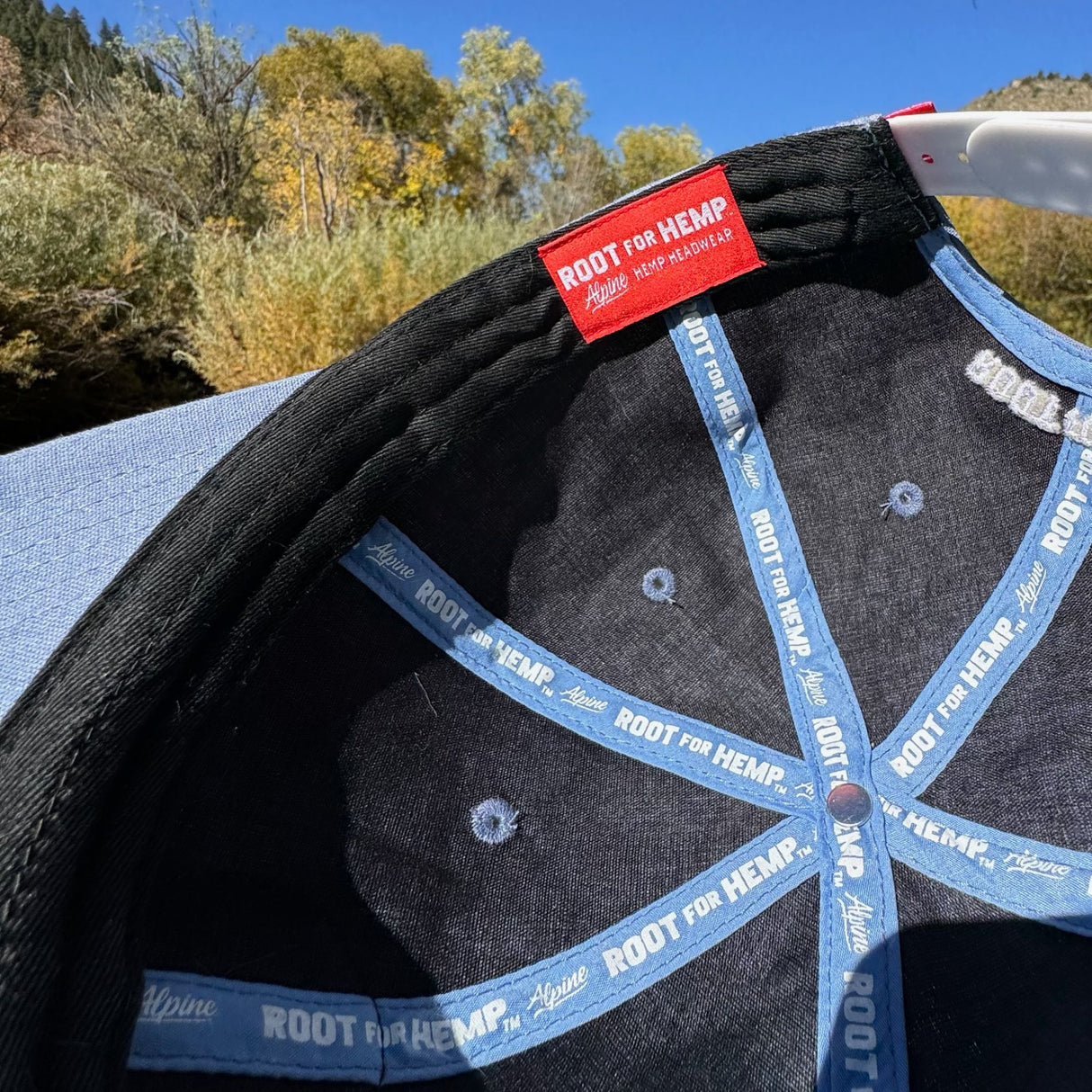 Inside view of the Blue hemp vote baseball hat - highlighting the branded root for hemp on blue ribbon on the inside seams, inside of hat is a charcoal color.  Pictured outside with fall trees in the background. 