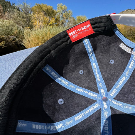 Inside view of the Blue hemp vote baseball hat - highlighting the branded root for hemp on blue ribbon on the inside seams, inside of hat is a charcoal color.  Pictured outside with fall trees in the background. 