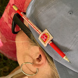 Closeup view of the pencil holder on the blue root for hemp vote hat in use.  Pencil is red with hemp educational messaging.  The hat is on a model with blond hair, wearing black glasses and silver hoop earrings.  