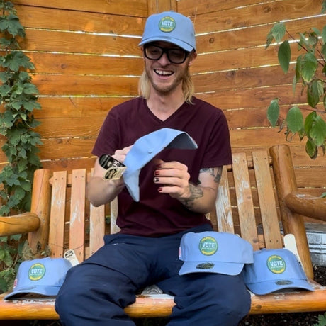 Person sitting on a wooden bench with a few blue hemp vote hats on their lap and on in their hands.  Person is wearing blue pants, and a maroon v-neck t-shirt and the hemp vote hat.  