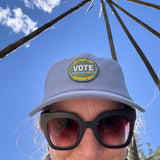 Front view of the hemp vote hat on a model.  Hat is blue with the words root for with a green embroidered patch that says "Vote Like Your Leaf Depends on it".  Model is wearing black sunglasses, background is blue sky with a few clouds.