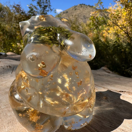 Hand poured resin figurine of a full bodied woman, with hemp buds at heart-center and gold flake throughout.  Figurine is sitting on a wooden table with a mountain/tree scene in the background.