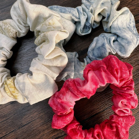 Closeup view of three hemp eco print scrunchies - left is natural color, right is blue color, bottom is red color.  Pictured on dark wooden table.