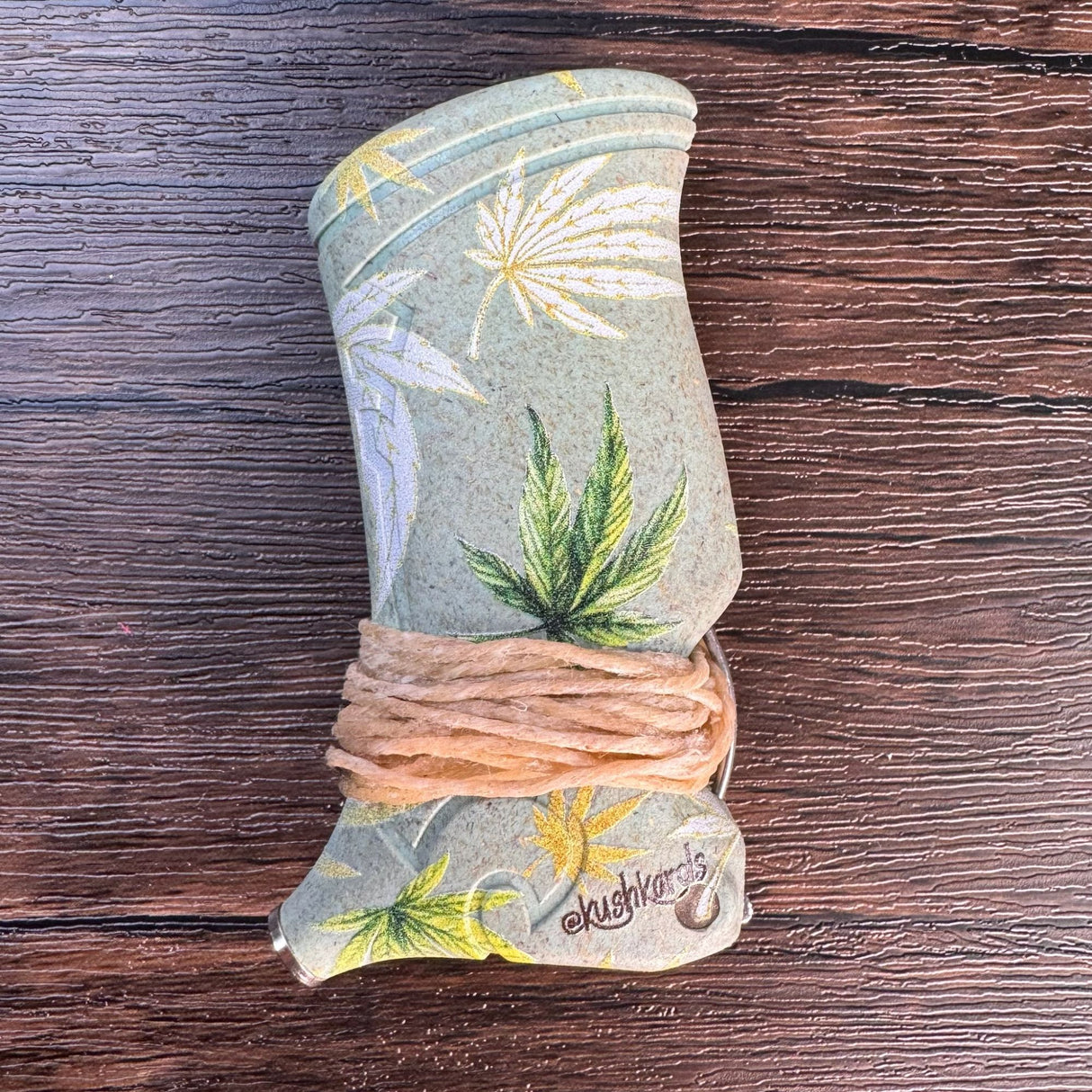 Closeup view of a toker poker that is light green with hemp leaves printed in yellow, green and white.  Toker is wrapped with hemp wick and pictured on a wooden surface. 