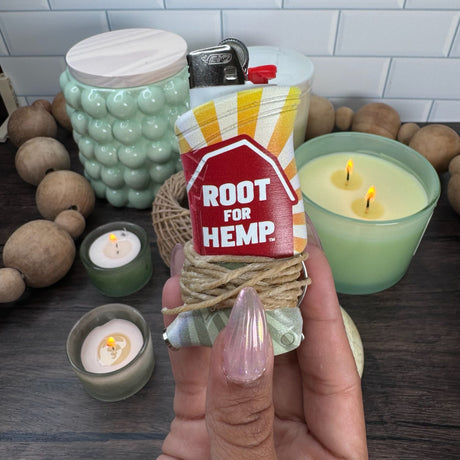 Closeup view of a toker poker with the words "root for hemp" in white text on a red barn.  There are yellow sunburst and green fields also in the design.  Toker is wrapped with hemp wick and pictured with candles and beads in the background.